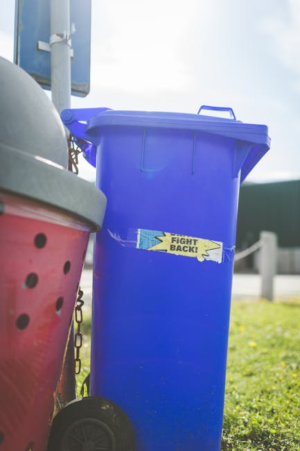 A green, metal recycling bin designated for old clothing collection is situated outdoors against a backdrop of dense foliage and wooden fencing. The bin features an open rectangular slot at the top, l